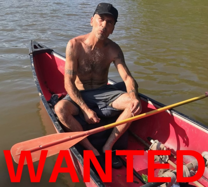 topless man with tattoos wearing baseball cap and shorts, sitting in a boat, with the word "WANTED" printed at the bottom