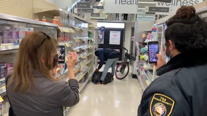 A shoplifter with a bike fills a garbage bag with goods as a woman and a security guard film the incident on their phones