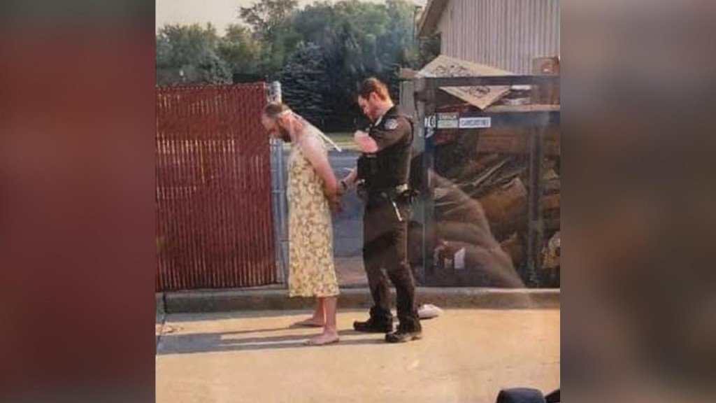 Andrew Loudon wearing a yeloow pattern dress being handcuffed by a police officer